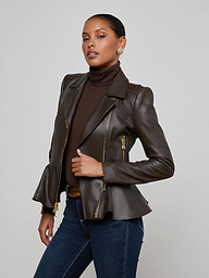 Wearing the Lyric Leather Peplum Jacket over a dark turtleneck, a woman stands confidently with her hand on her waist, styled with jeans and earrings as she poses against a plain light background.