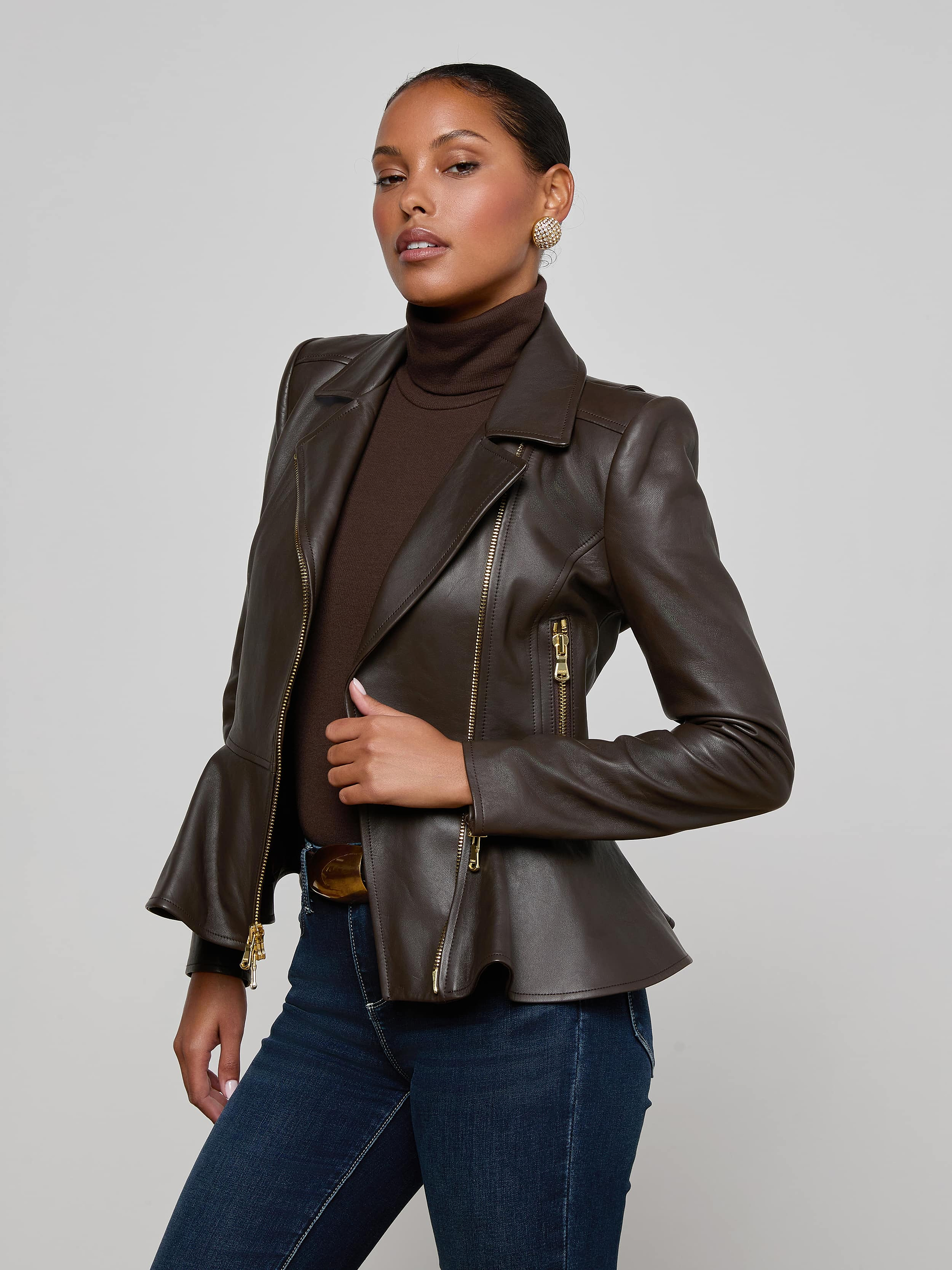 Wearing the Lyric Leather Peplum Jacket over a dark turtleneck, a woman stands confidently with her hand on her waist, styled with jeans and earrings as she poses against a plain light background.