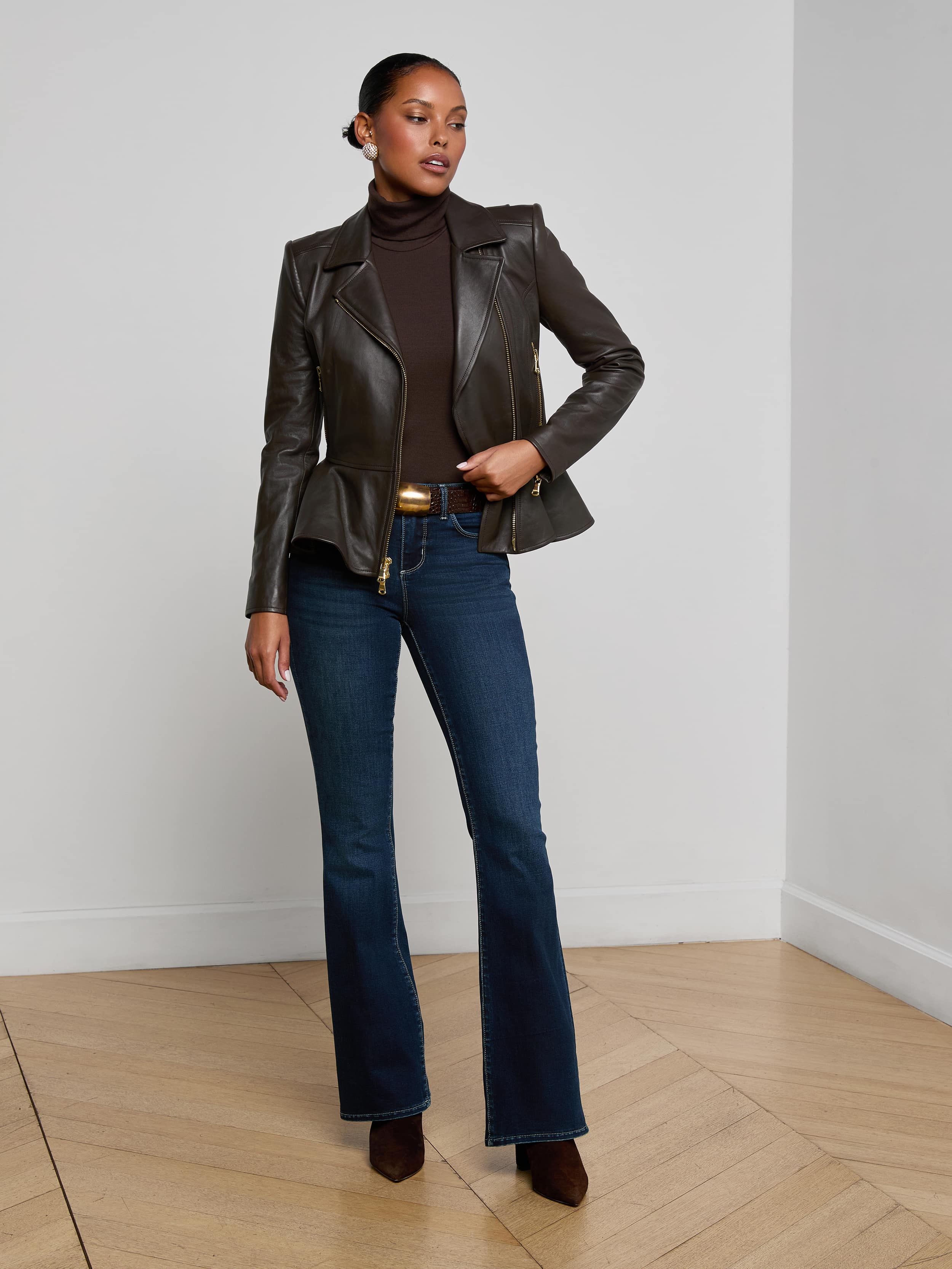 A woman stands against a plain light background, wearing the Lyric Leather Peplum Jacket in black, paired with a dark turtleneck, blue flared jeans, brown shoes, and gold hoop earrings. She has one hand in her pocket and looks confidently ahead.