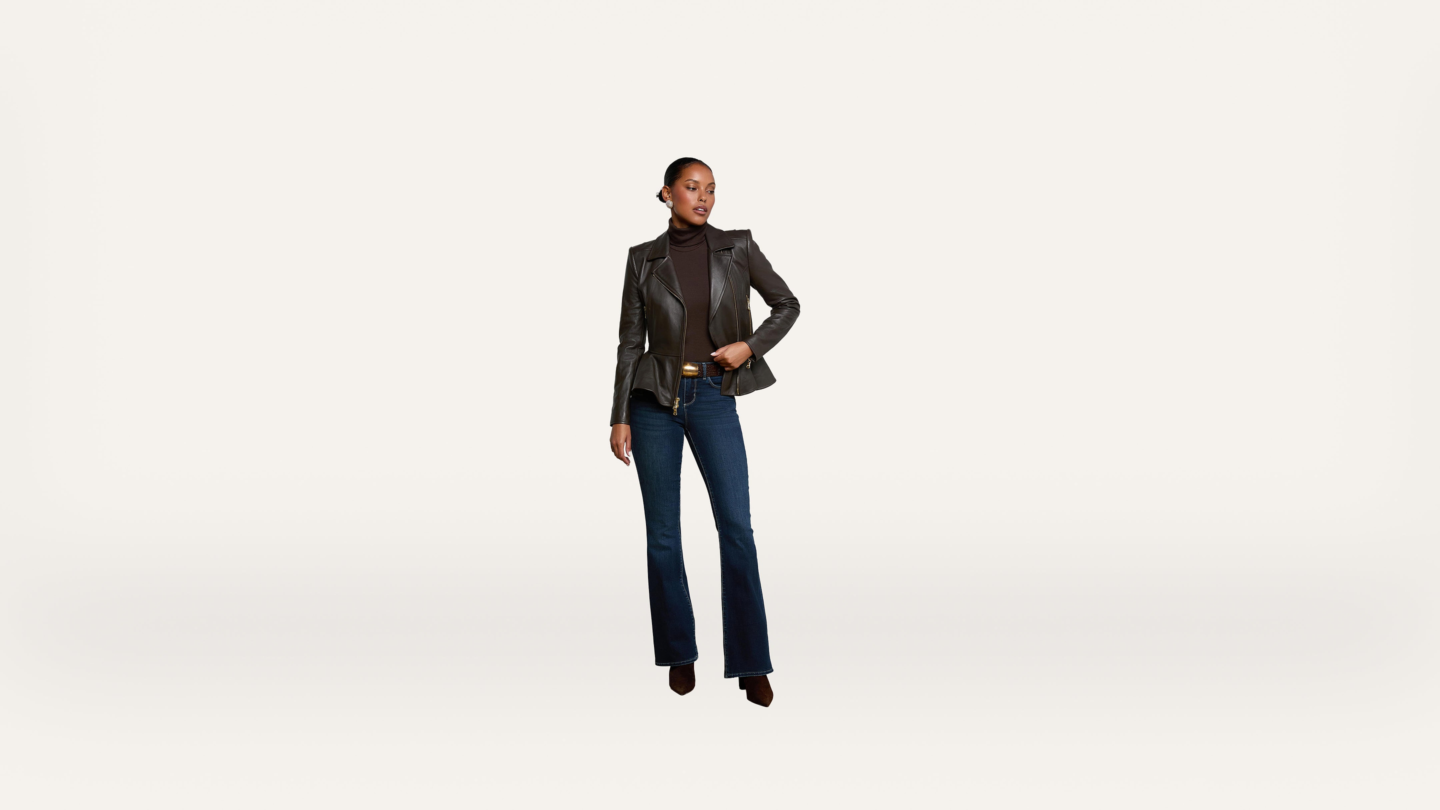A woman wearing a black leather blazer, dark turtleneck, blue flared jeans, and heeled boots stands confidently against a plain white background.