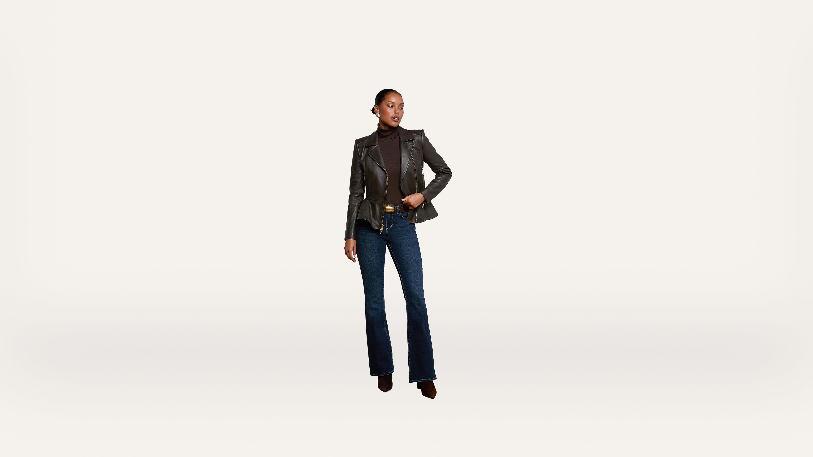 A woman wearing a black leather blazer, dark turtleneck, blue flared jeans, and heeled boots stands confidently against a plain white background.