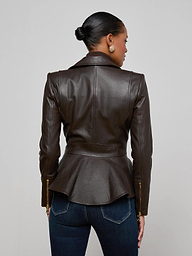 A woman stands facing away, wearing the Lyric Leather Peplum Jacket—a feminine dark brown leather piece with a peplum hem—paired with blue jeans. Her hair is styled in a neat bun, set against a plain off-white background.