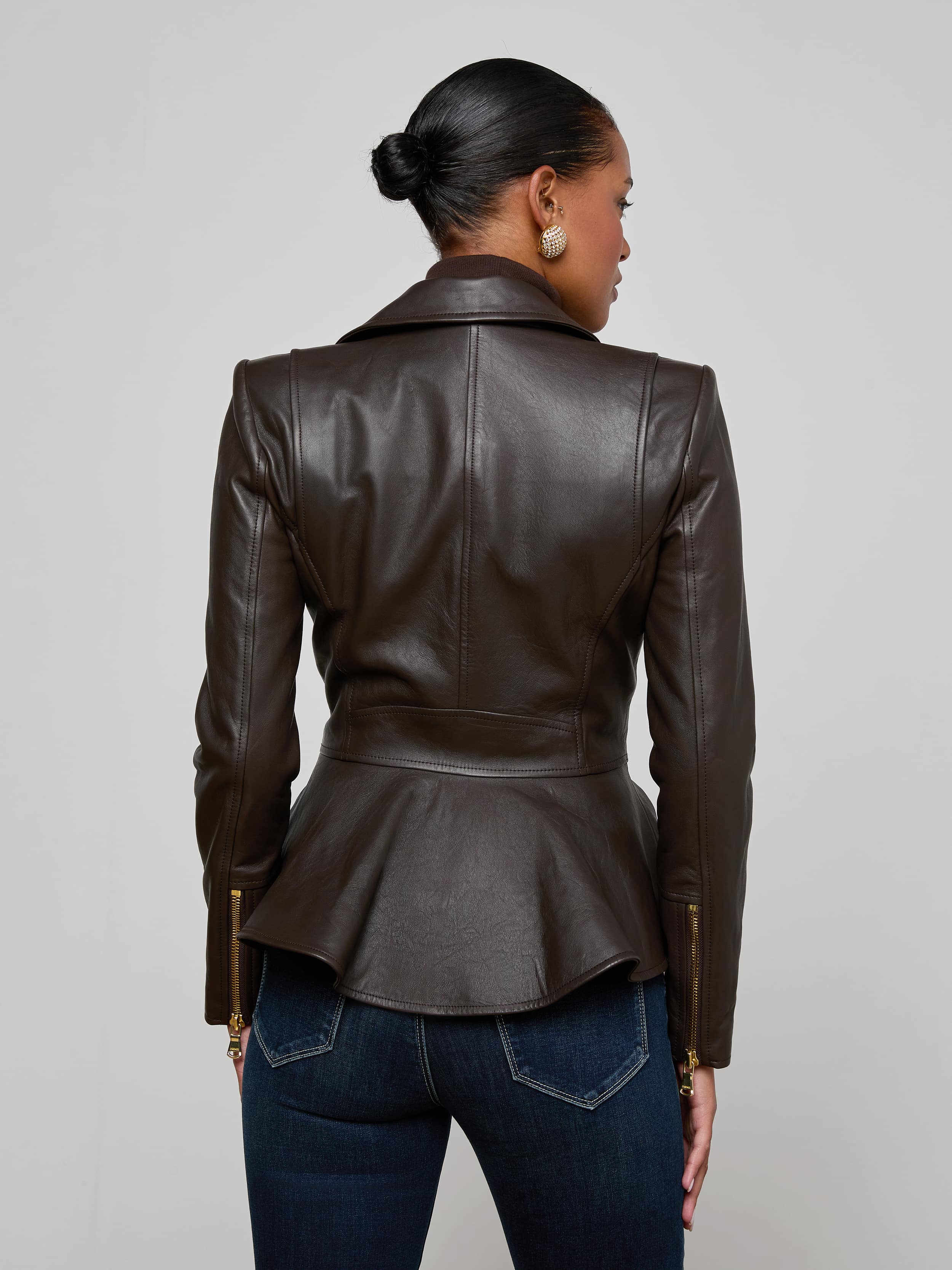 A woman stands facing away, wearing the Lyric Leather Peplum Jacket—a feminine dark brown leather piece with a peplum hem—paired with blue jeans. Her hair is styled in a neat bun, set against a plain off-white background.