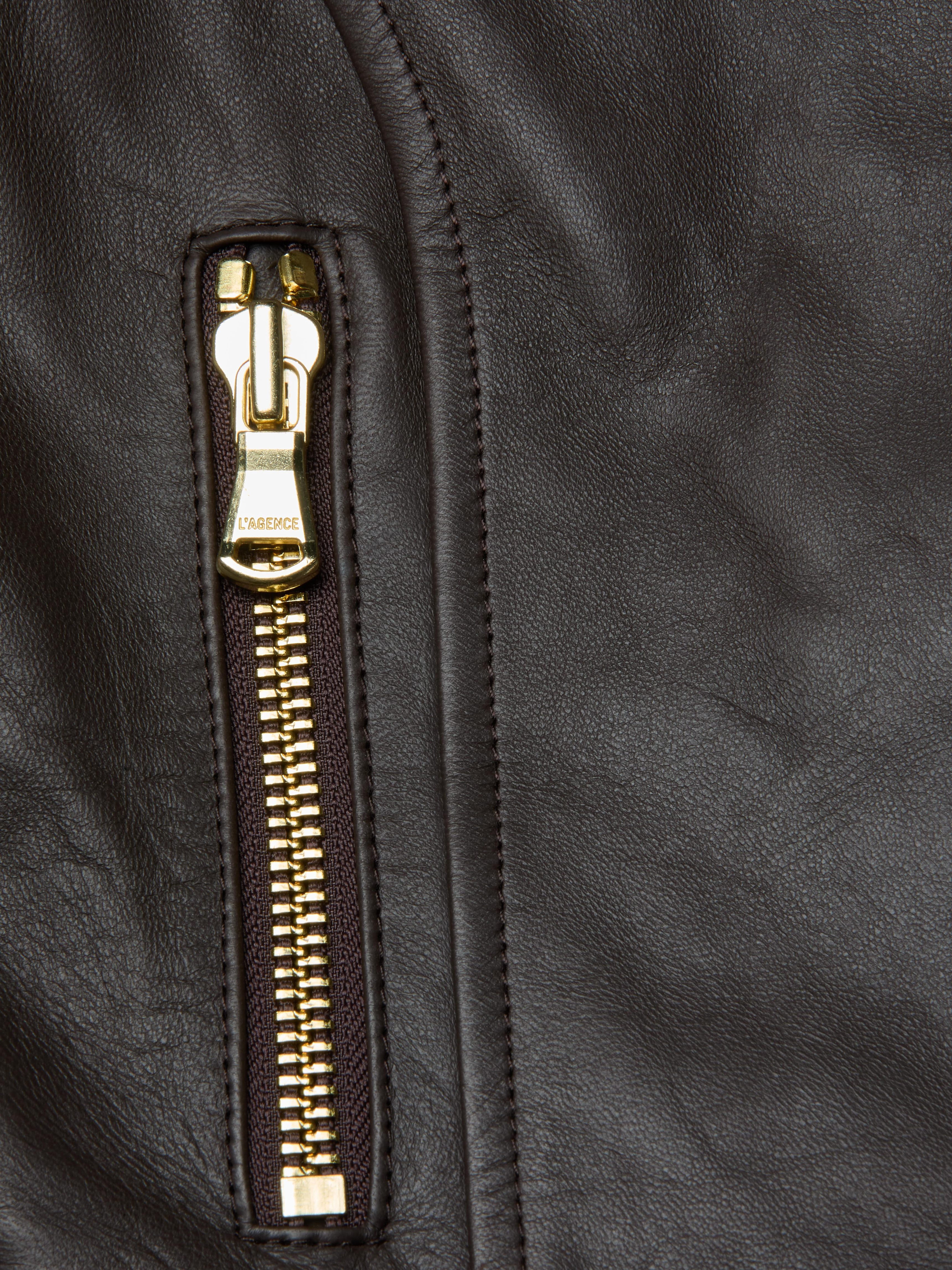 A close-up of a gold zipper on dark brown leather, highlighting the textured and smooth surfaces of the Lyric Leather Peplum Jacket. The zipper pull is labeled LAGENCE, emphasizing the quality details characteristic of this feminine silhouette.