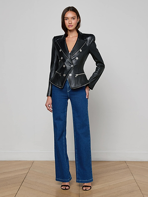 A woman stands against a light gray wall wearing the L'AGENCE Ottilie Leather Blazer, blue high-waisted wide-leg jeans, and black open-toe heels. With straight hair and a neutral expression, she epitomizes effortless power dressing.