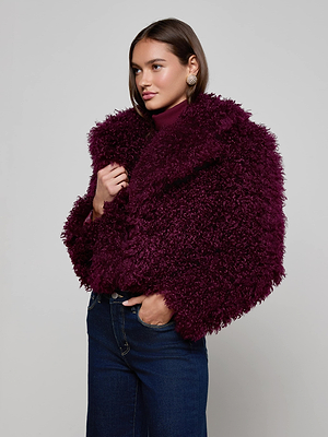 A woman with straight brown hair wears the L'AGENCE Aura Cropped Faux Fur Jacket in burgundy over a matching turtleneck and dark blue jeans, standing against a light gray background.