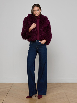 A woman poses indoors, touching her L'AGENCE Aura Cropped Faux Fur Jacket in deep burgundy with a curly texture, styled with a matching turtleneck, dark blue high-waisted wide-leg jeans, and dark purple heels.