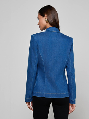 A woman with straight brown hair, gold hoop earrings, and black pants models the L'AGENCE Baldwin Blazer in indigo wash, standing with her back to the camera against a plain light gray background.