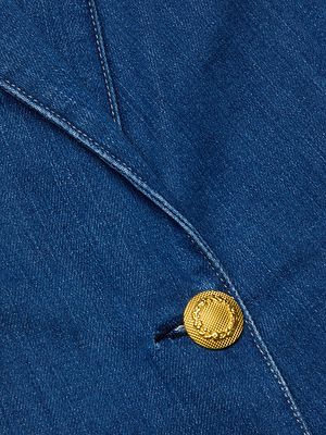 Close-up of the Baldwin Blazer by L'AGENCE, featuring blue indigo wash denim, a gold textured button, and visible white stitching along the fabric edge.