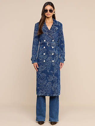 A person wearing sunglasses, the Bradley Trench Coat—a long blue floral jacquard patterned coat with buttons and a belt—along with blue pants and dark shoes stands against a beige background.
