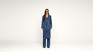 Woman standing against a plain background, wearing sunglasses, a long blue patterned coat with buttons, and flared blue jeans, facing forward with hands by her sides.