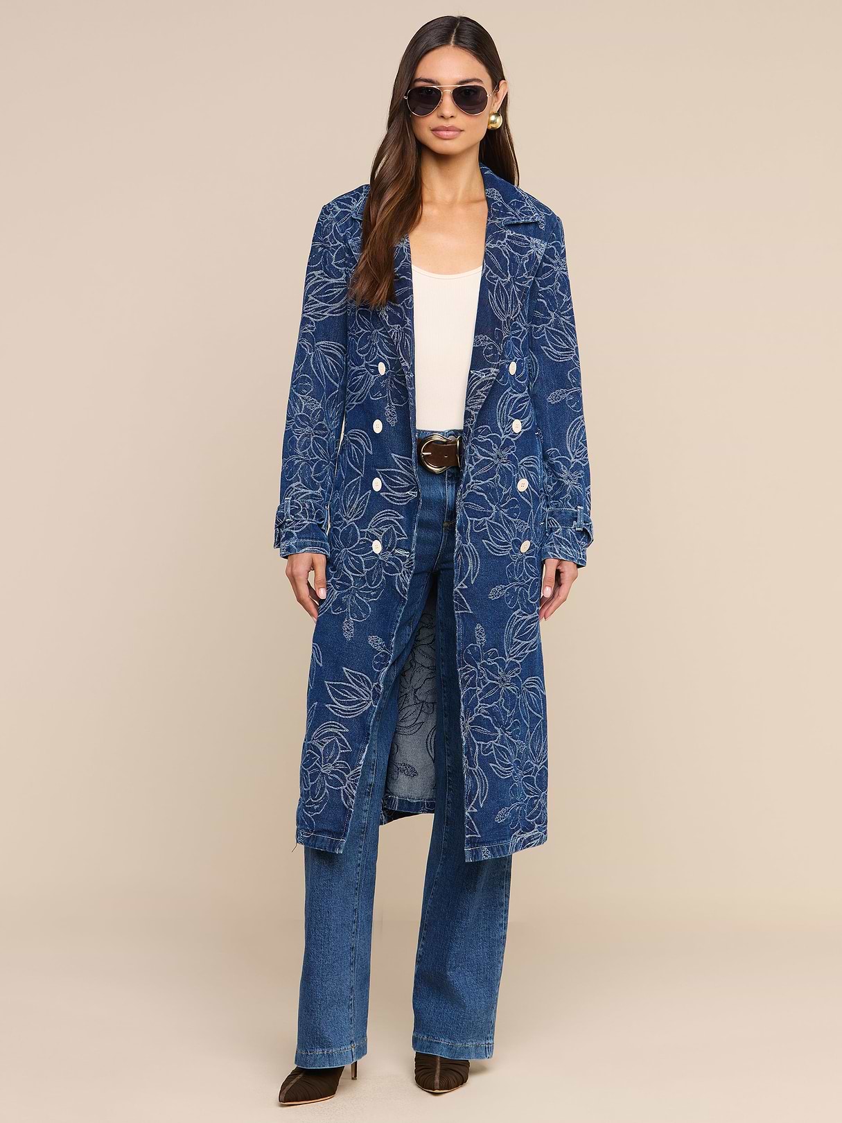 A woman wearing sunglasses and the Bradley Trench Coat—a long blue floral jacquard style—over a white top, blue jeans, and dark shoes stands against a plain white background.