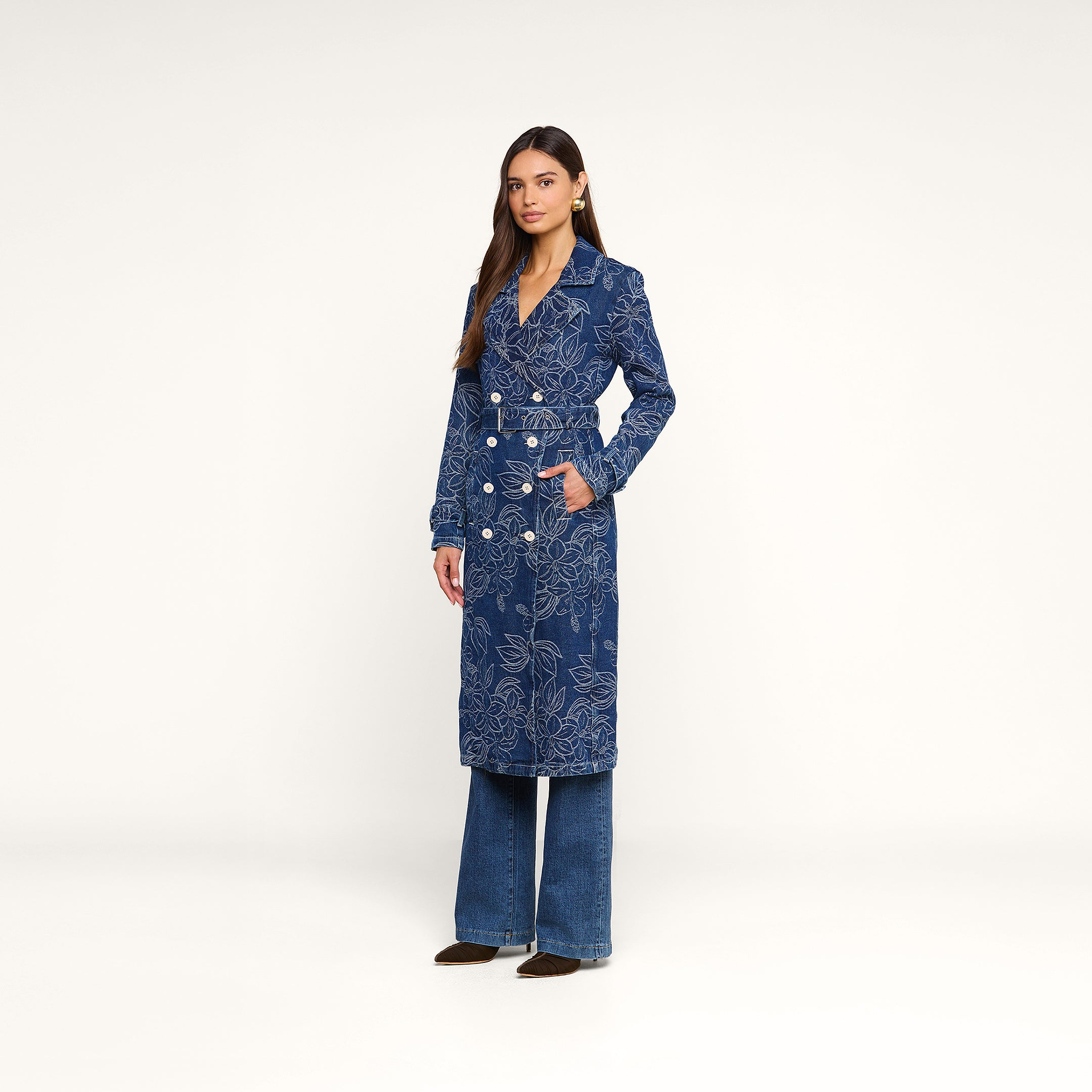 A woman stands in the Bradley Trench Coat—a blue, floral jacquard double-breasted style with white buttons—paired with wide-leg jeans and brown shoes, set against a plain white background.
