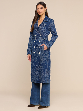 A woman stands in the Bradley Trench Coat, a double-breasted blue denim piece with a white floral jacquard pattern, paired with dark blue wide-leg jeans and brown shoes, posing against a plain beige background.