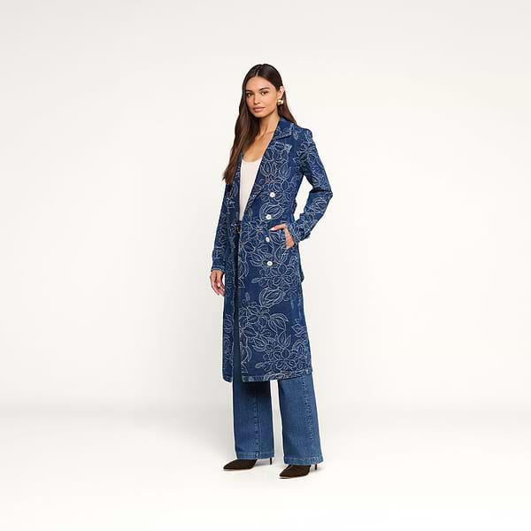 A woman stands against a plain white background, wearing the Bradley Trench Coat—a long, blue floral jacquard design—paired with blue jeans and brown shoes. With one hand in her pocket, she looks at the camera.