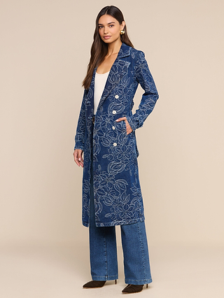 A woman stands with one hand in her pocket against a beige background, wearing the Bradley Trench Coat—a long, blue floral jacquard denim trench coat with a double-breasted design—layered over a white top and blue jeans.