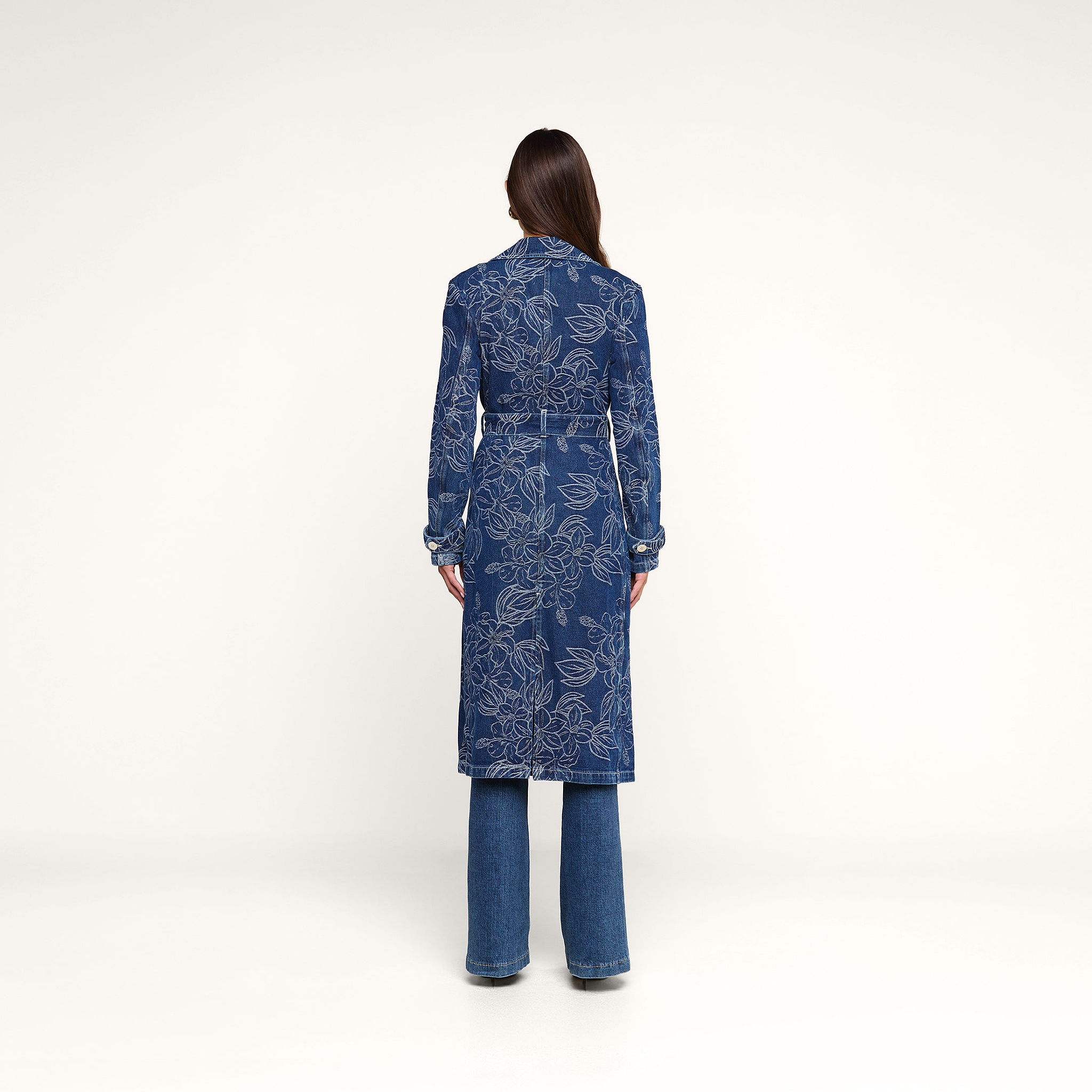 A person stands facing away, wearing the Bradley Trench Coat—a long blue floral jacquard trench coat—and blue jeans, against a plain white background.
