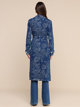A woman with long brown hair stands facing away against a plain beige background, wearing the Bradley Trench Coat—a long, blue, floral jacquard denim trench coat—paired with matching denim pants.