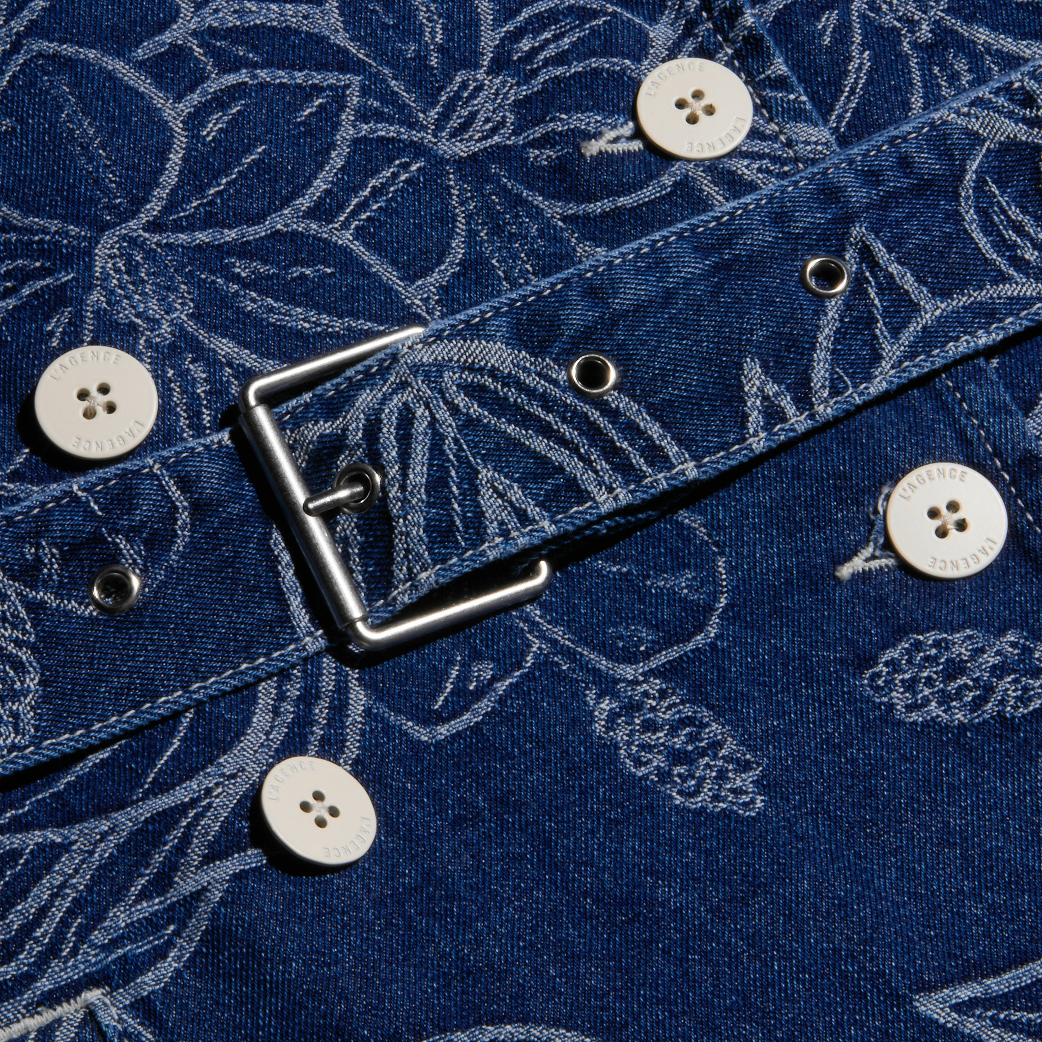 Close-up of the blue floral jacquard denim fabric with white buttons, metal grommets, and a square metal buckle from the Bradley Trench Coat.