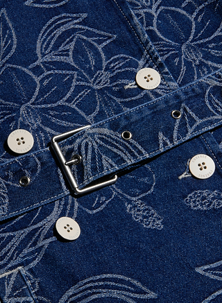 Close-up of the Bradley Trench Coat in blue denim fabric, featuring white floral jacquard patterns, white buttons, and a silver metal buckle. Visible stitching and buttonholes emphasize the texture and design details of this double-breasted trench coat.