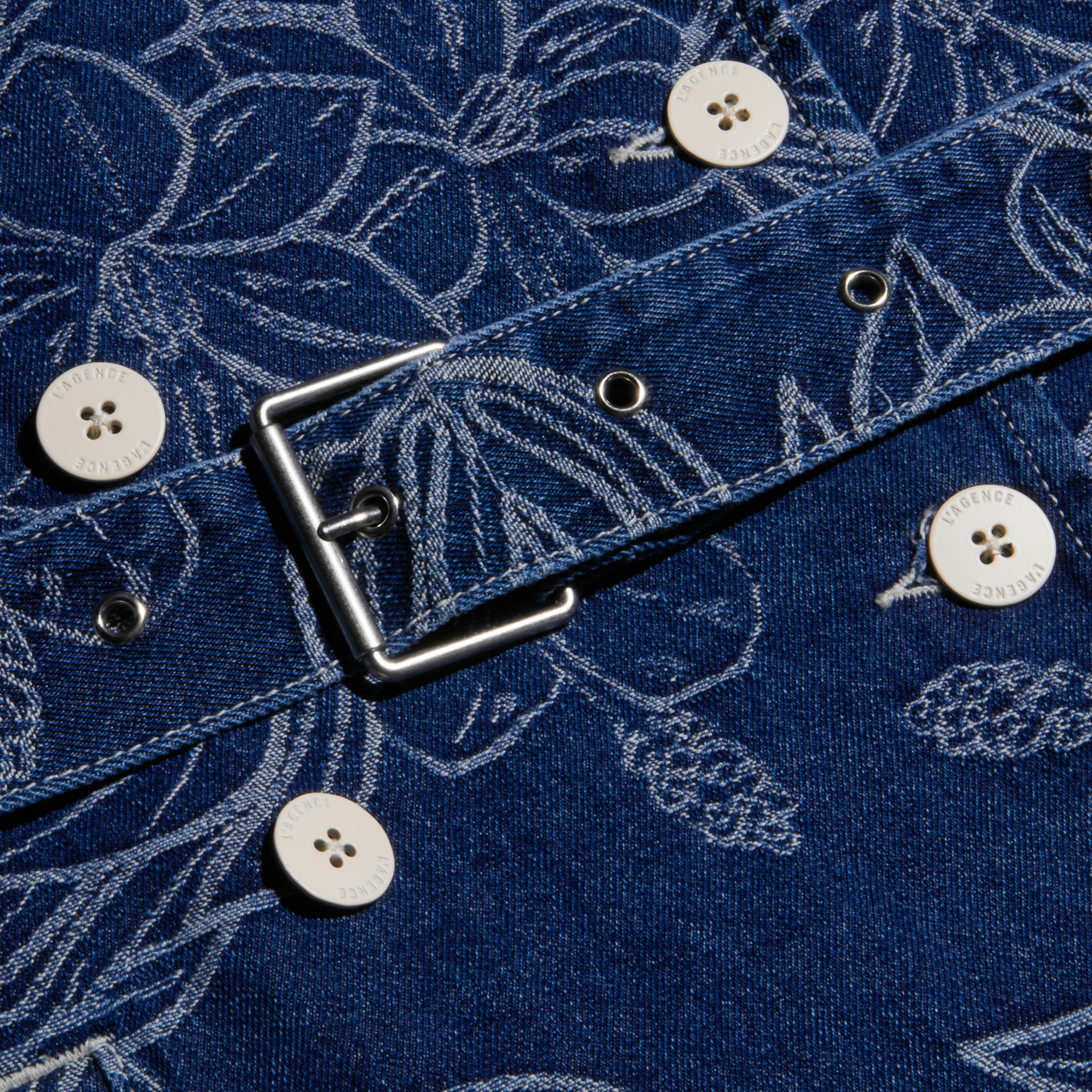 Close-up of the blue floral jacquard denim fabric with white buttons, metal grommets, and a square metal buckle from the Bradley Trench Coat.