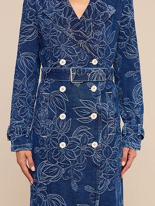 A person wearing the Bradley Trench Coat—a blue floral jacquard denim trench coat featuring white floral patterns, white buttons, and a matching belt. Only the torso, arms, and hands are visible against a beige background.
