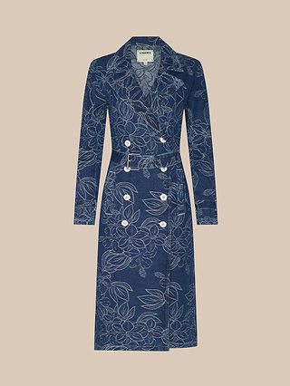 The Bradley Trench Coat features a long blue denim design with a belted waist, white buttons, and an outlined floral jacquard pattern throughout, showcased against a neutral beige background.