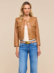 A woman with blonde hair stands against a beige background, looking directly at the camera. She is wearing the Deidra Cropped Leather Jacket—a tan, collarless leather jacket featuring large gold buttons—styled with a beige top, blue jeans, and a tan belt with a gold buckle.
