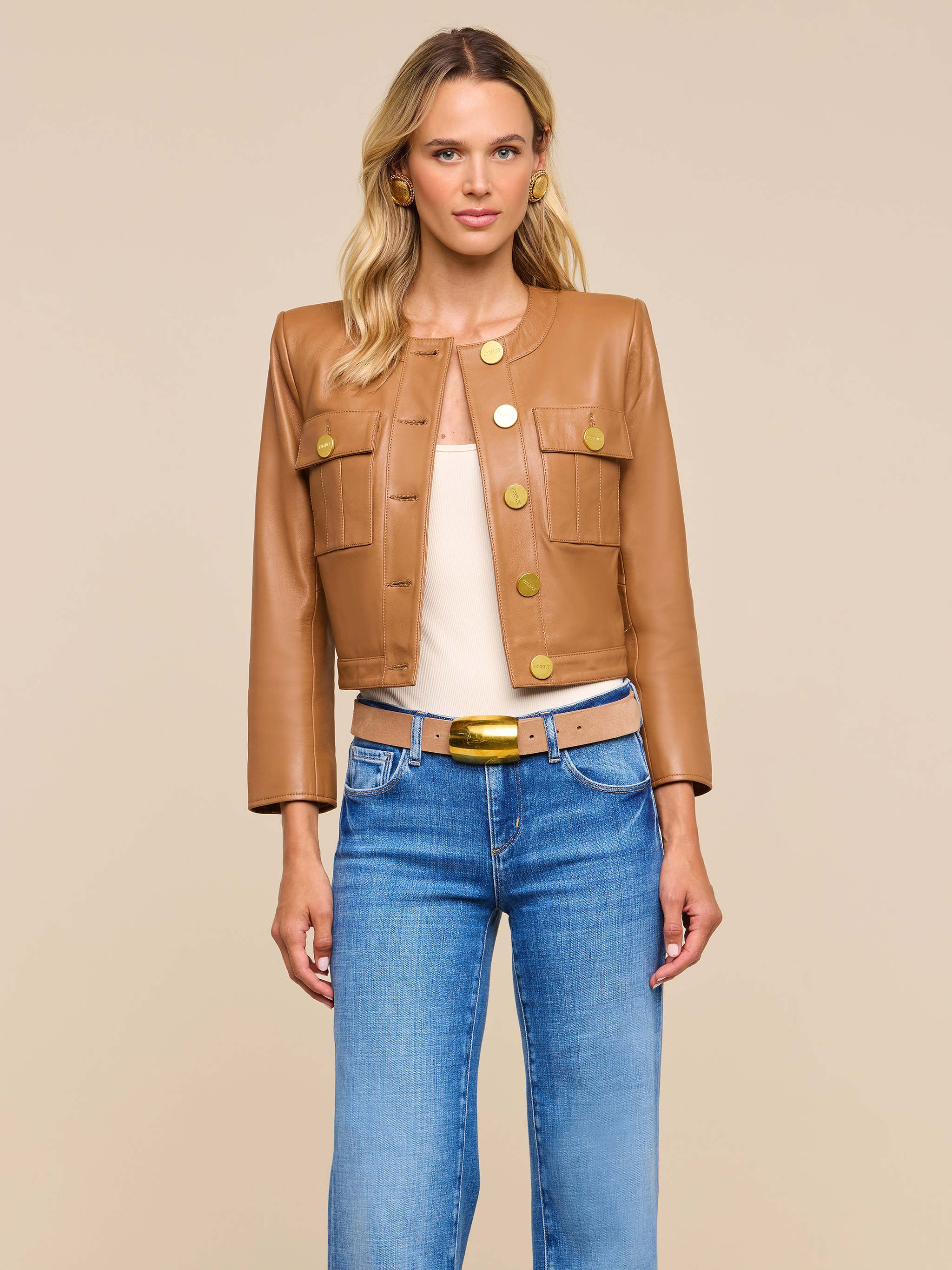 A woman with blonde hair stands against a beige background, looking directly at the camera. She is wearing the Deidra Cropped Leather Jacket—a tan, collarless leather jacket featuring large gold buttons—styled with a beige top, blue jeans, and a tan belt with a gold buckle.