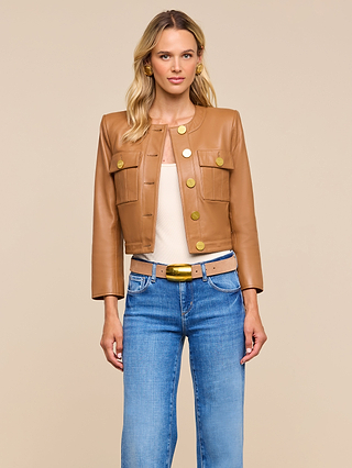 A woman with blonde hair stands against a beige background, looking directly at the camera. She is wearing the Deidra Cropped Leather Jacket—a tan, collarless leather jacket featuring large gold buttons—styled with a beige top, blue jeans, and a tan belt with a gold buckle.