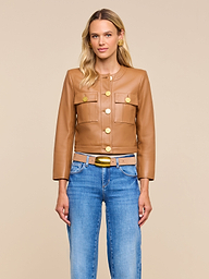 A woman wearing the Deidra Cropped Leather Jacket—tan, collarless, with gold buttons—paired with blue jeans stands against a beige background, looking forward with a neutral expression.
