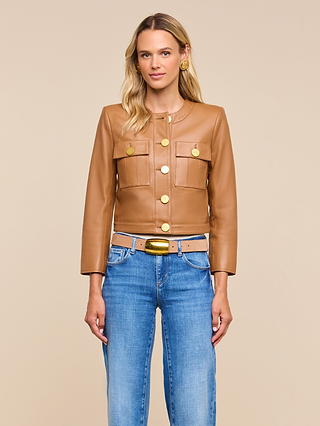 A woman wearing the Deidra Cropped Leather Jacket—tan, collarless, with gold buttons—paired with blue jeans stands against a beige background, looking forward with a neutral expression.