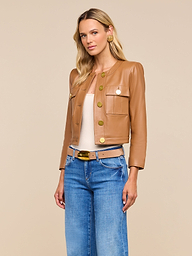 Woman wearing the Deidra Cropped Leather Jacket in tan, featuring a collarless design and large gold buttons, paired with a beige top, blue jeans, and a beige belt, standing against a plain beige background.