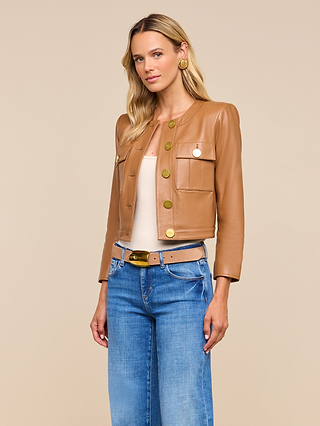 Woman wearing the Deidra Cropped Leather Jacket in tan, featuring a collarless design and large gold buttons, paired with a beige top, blue jeans, and a beige belt, standing against a plain beige background.