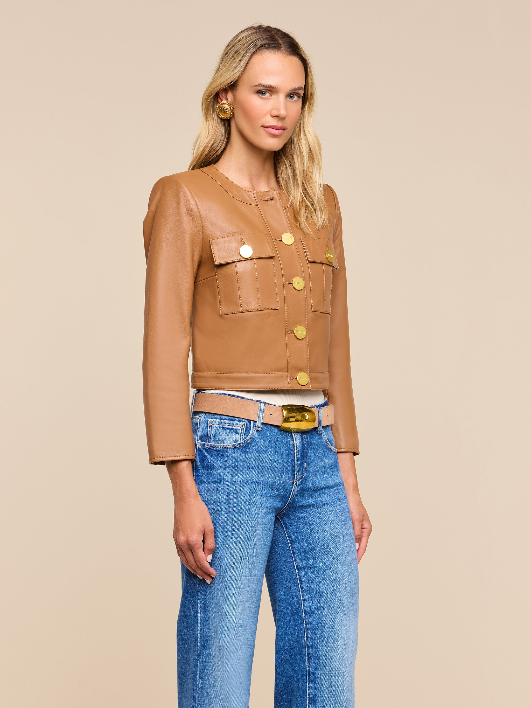 A woman wearing the Deidra Cropped Leather Jacket—featuring a tan hue, large gold buttons, and front pockets—along with blue jeans, a white belt, and gold earrings stands against a beige background.