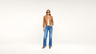 Woman wearing sunglasses, a tan jacket, white top, blue wide-leg jeans, and brown heels stands confidently against a plain white background.