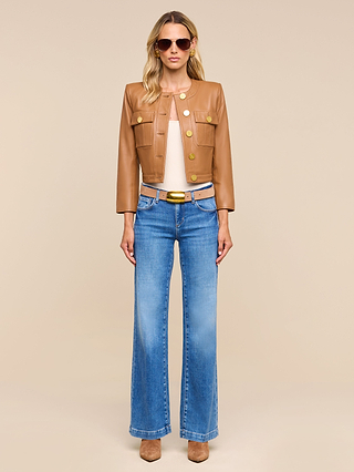 Wearing sunglasses, the person stands against a beige background facing forward with a neutral expression, dressed in the Deidra Cropped Leather Jacket—a tan, collarless and cropped leather piece—paired with a light beige top, blue flared jeans, and tan shoes.
