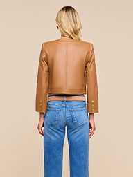 A woman with blonde hair stands facing away, wearing the Deidra Cropped Leather Jacket—a tan, collarless leather jacket with gold buttons on the sleeves—paired with blue jeans featuring visible back pockets, set against a beige background.
