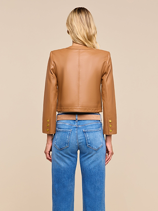 A woman with blonde hair stands facing away, wearing the Deidra Cropped Leather Jacket—a tan, collarless leather jacket with gold buttons on the sleeves—paired with blue jeans featuring visible back pockets, set against a beige background.