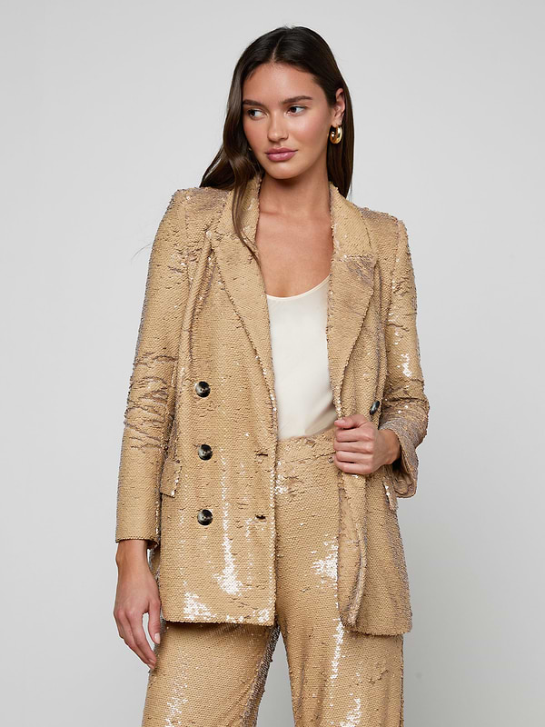 A woman stands against a light background wearing the Cambrie Oversized Sequin Blazer—a shiny gold, oversized sequin piece styled with a white top underneath. She has long brown hair and gold hoop earrings, looking slightly to the side with a confident expression—a chic nod to Spring 2026 fashion.