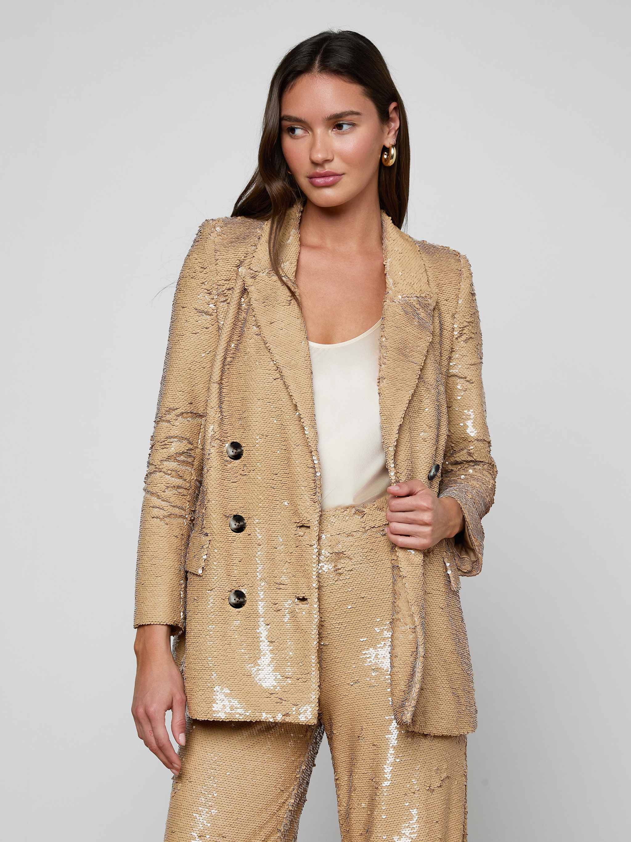 A woman stands against a light background wearing the Cambrie Oversized Sequin Blazer—a shiny gold, oversized sequin piece styled with a white top underneath. She has long brown hair and gold hoop earrings, looking slightly to the side with a confident expression—a chic nod to Spring 2026 fashion.