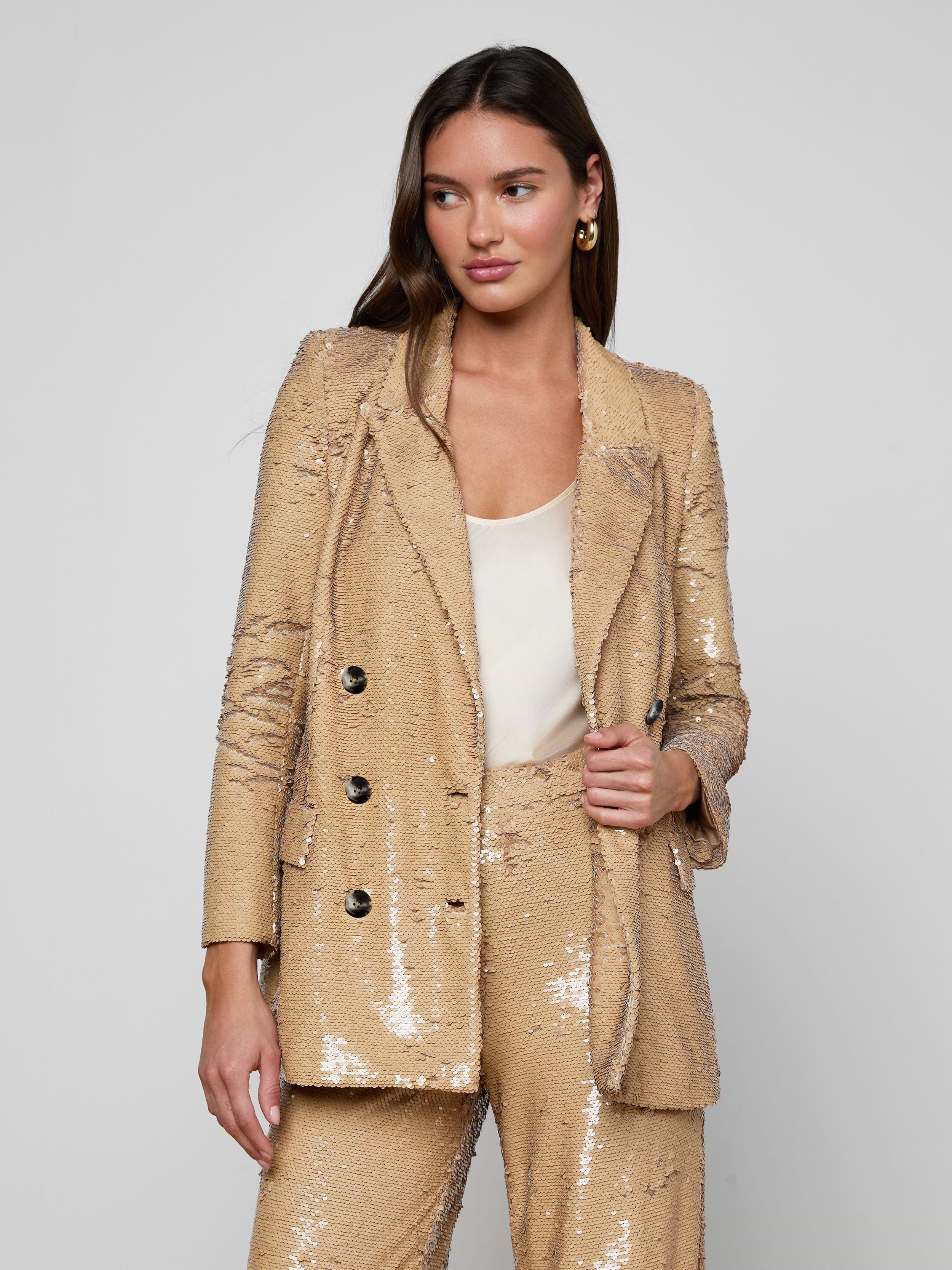 A woman stands against a light background wearing the Cambrie Oversized Sequin Blazer—a shiny gold, oversized sequin piece styled with a white top underneath. She has long brown hair and gold hoop earrings, looking slightly to the side with a confident expression—a chic nod to Spring 2026 fashion.