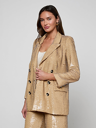 Dressed in the Cambrie Oversized Sequin Blazer with matching pants layered over a white top, the woman stands against a plain light background, gazing at the camera with a subtle smile—a perfect showcase of Spring 2026 fashion.