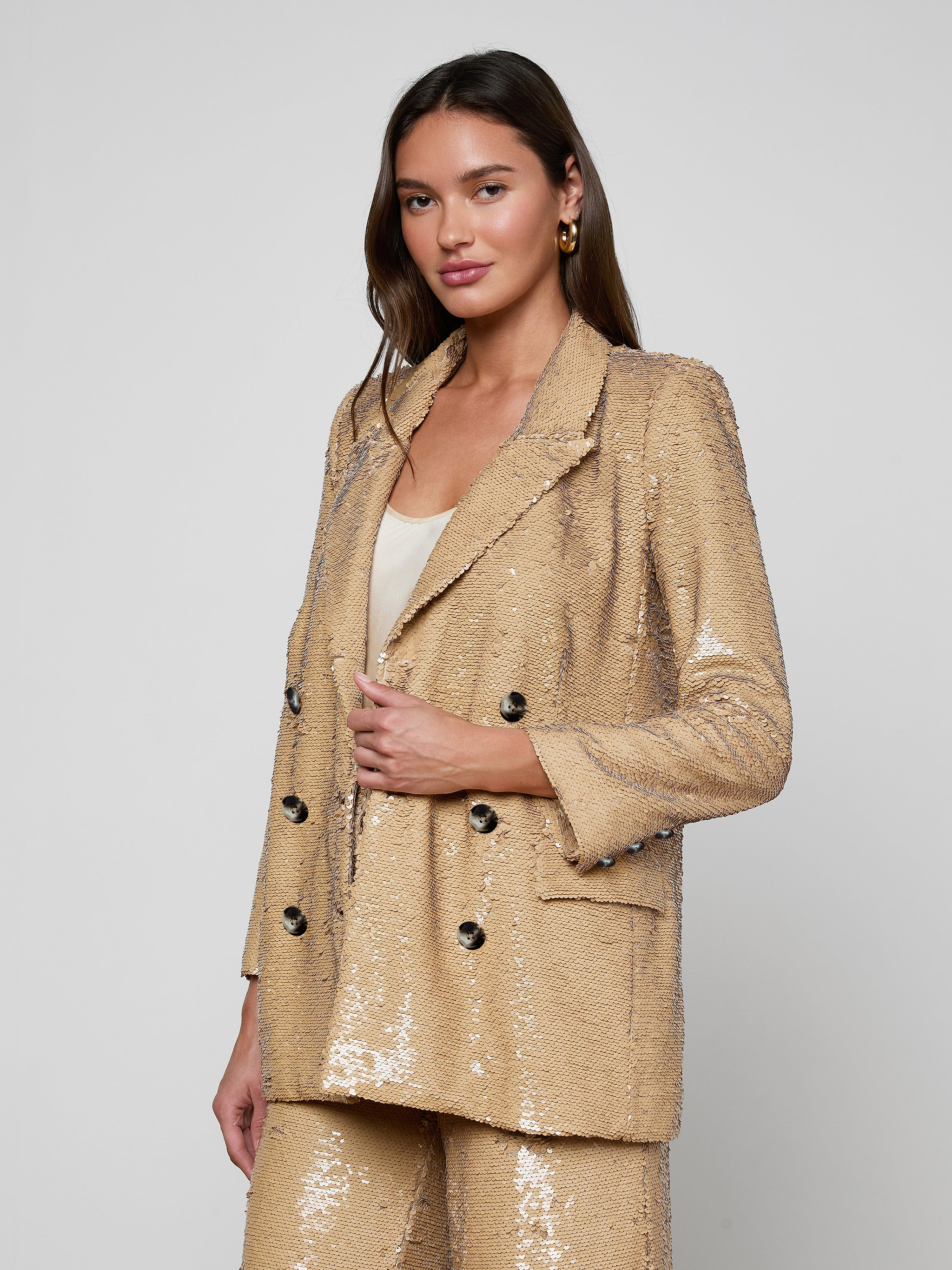 Dressed in the Cambrie Oversized Sequin Blazer with matching pants layered over a white top, the woman stands against a plain light background, gazing at the camera with a subtle smile—a perfect showcase of Spring 2026 fashion.