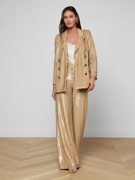 A woman stands against a plain white background, wearing the Cambrie Oversized Sequin Blazer in shimmering gold paired with matching wide-leg sequin pants and a white top underneath. This ensemble is perfect for making a bold statement in Spring 2026 fashion.