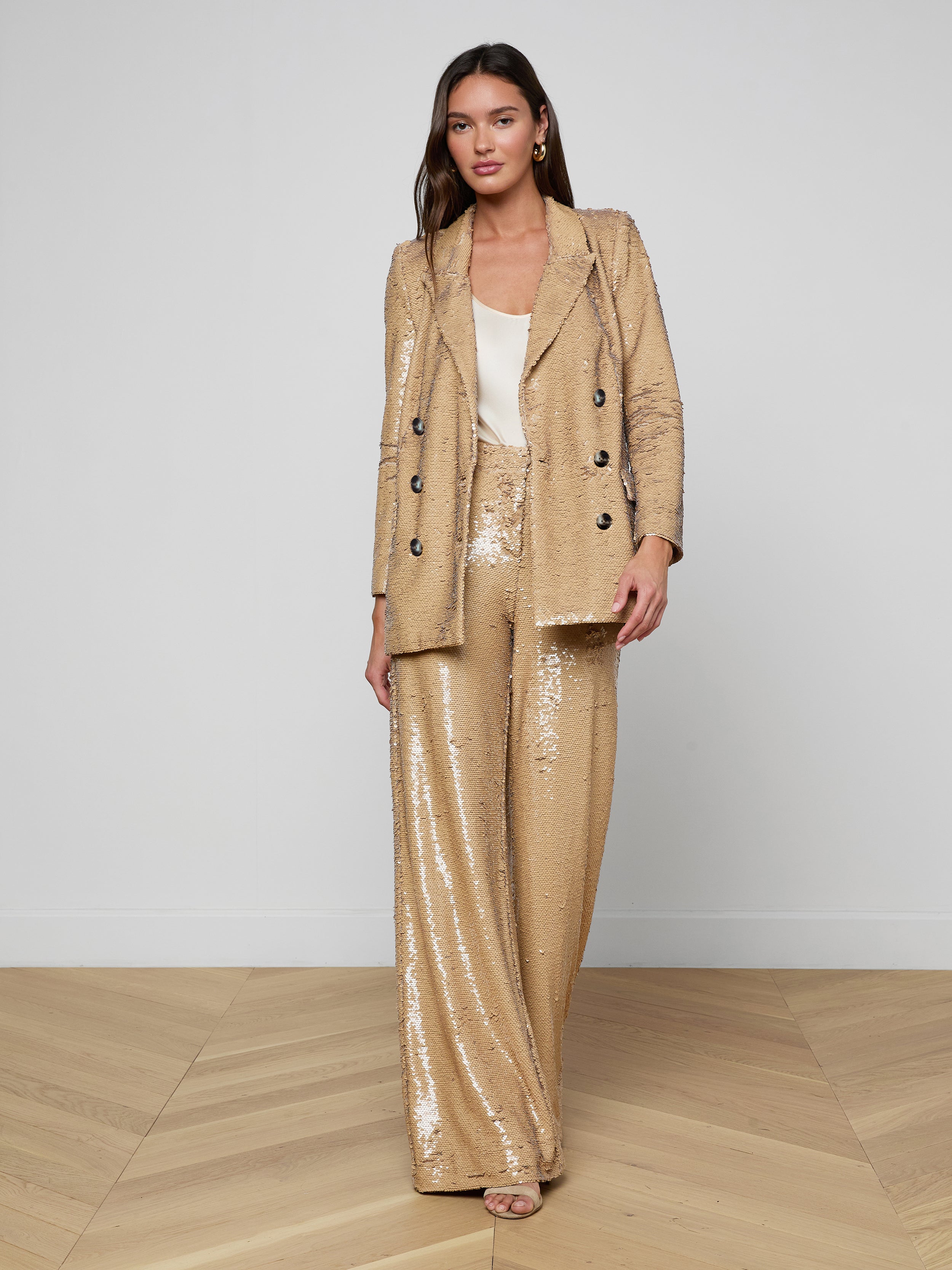 A woman stands against a plain white background, wearing the Cambrie Oversized Sequin Blazer in shimmering gold paired with matching wide-leg sequin pants and a white top underneath. This ensemble is perfect for making a bold statement in Spring 2026 fashion.