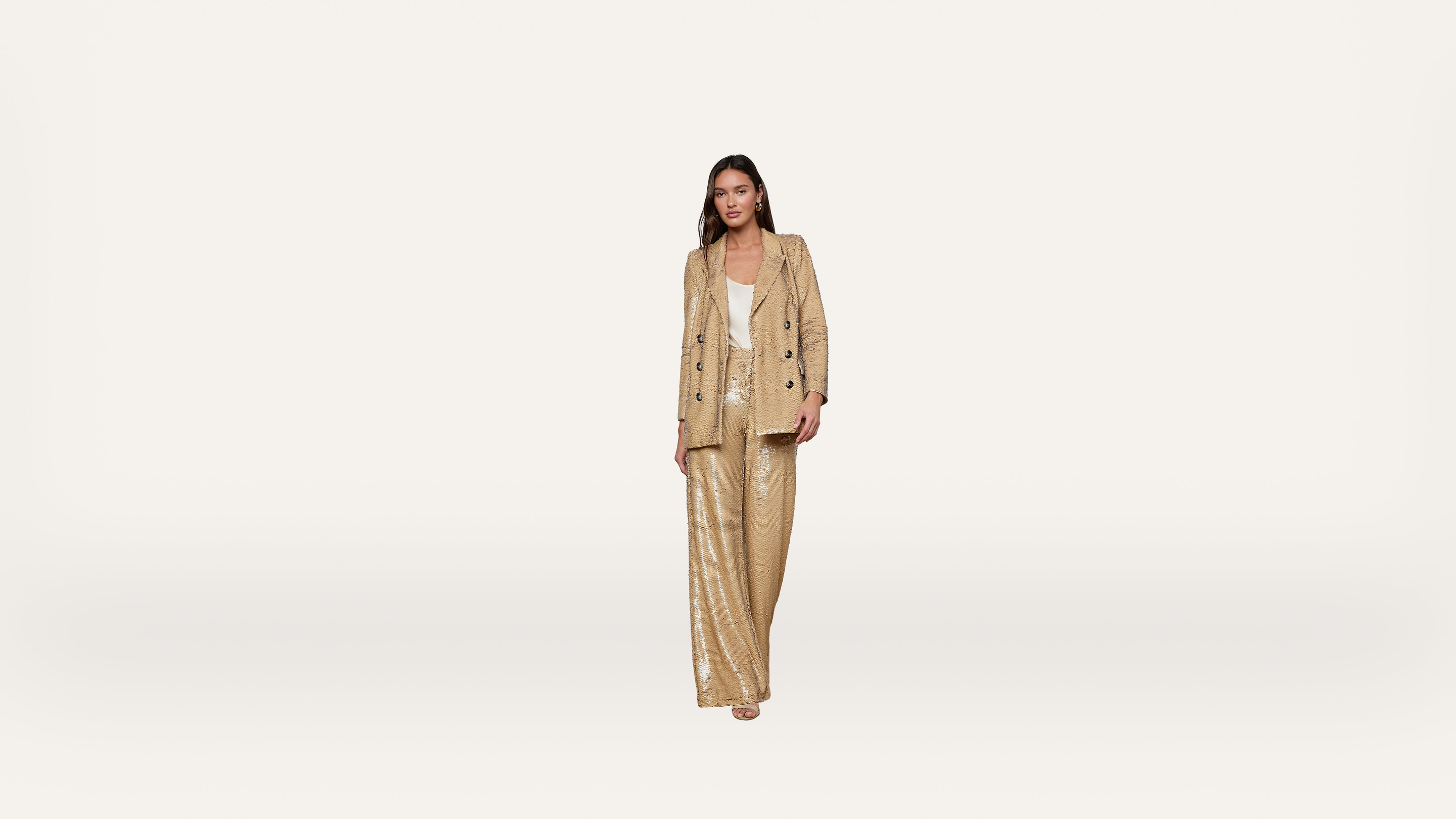 A woman stands against a plain white background wearing a beige textured pantsuit with a matching blazer and wide-leg trousers, paired with a white top and open-toed shoes.