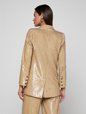 A woman faces away in the Cambrie Oversized Sequin Blazer and matching light gold pants, with straight brown hair and gold hoop earrings. Her left hand is at her side, highlighting Spring 2026 style against a plain, light background.