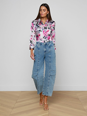 A woman stands indoors against a white wall and herringbone wood floor, wearing a shiny floral blouse, clear-strap heeled sandals, and light blue Reba Rodeo Jean barrel-leg denim.