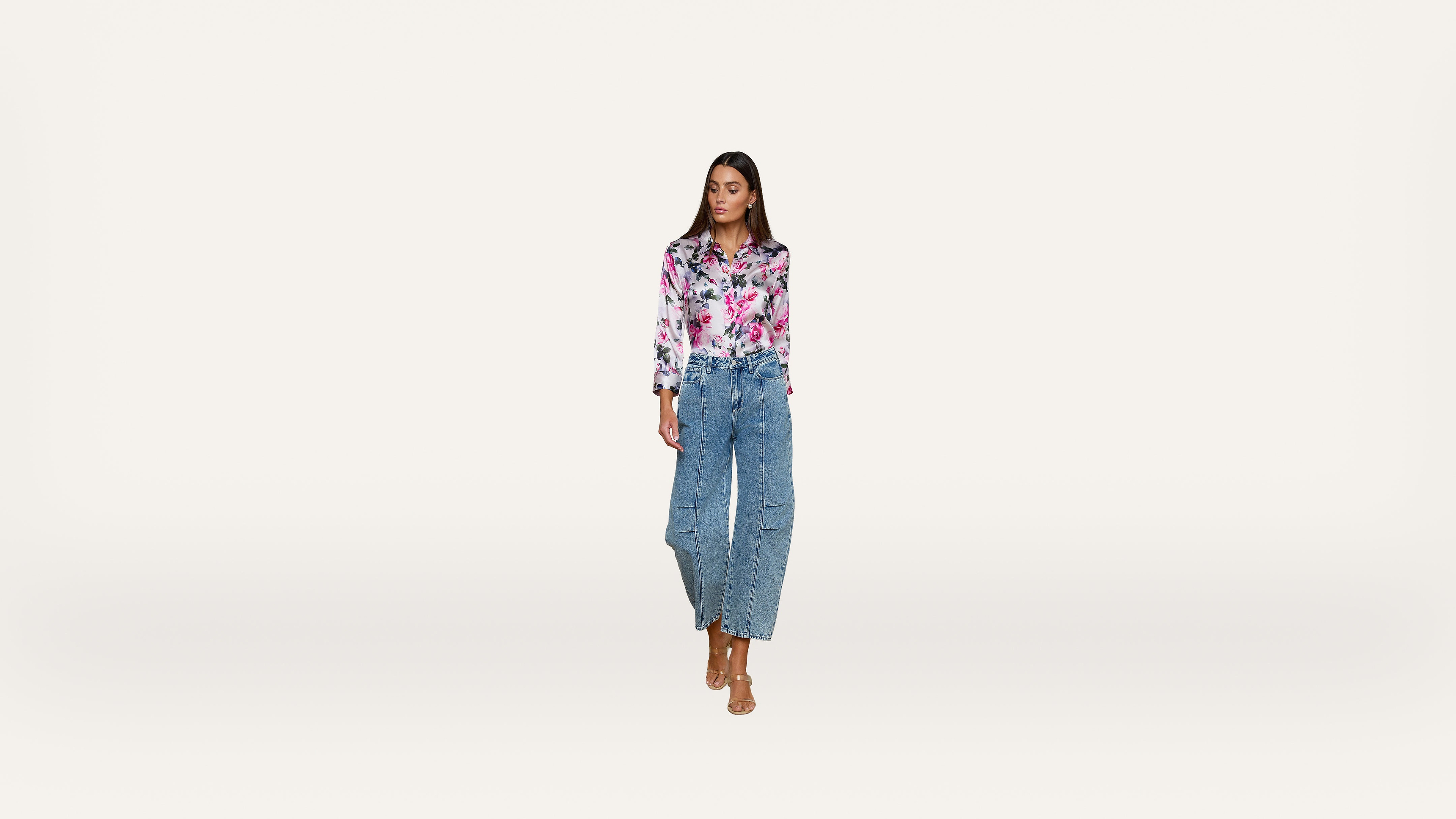 A woman stands against a plain white background, wearing a pink floral blouse, high-waisted blue jeans, and beige heeled sandals. She looks slightly to the side with her hands at her sides.